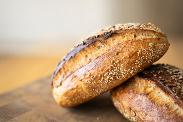 Two loaves of sourdough bread, freshly baked and still warm, are stacked on a wooden surface. The loaves are covered in seeds sesame and sunflower, adding a rustic and flavorful touch.
