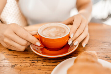 A woman is holding a red coffee cup with a white saucer. The woman is sitting at a table with a plate of croissants in front of her.