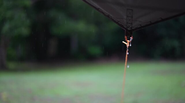 camping on rainy season with closeup rain water drips droplets to tip tarp and flow slow motion on fly sheet tent with carabiner paracord rope for holiday vacation picnic on green nature forest garden