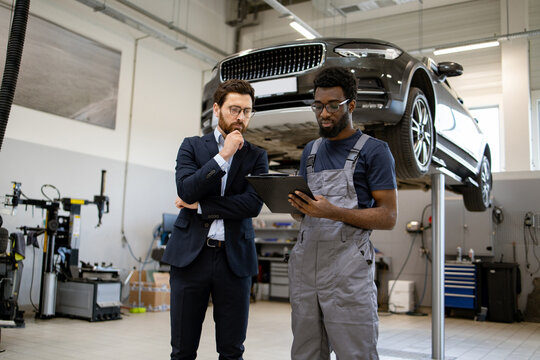Mechanic in uniform showing car repair details to businessman in suit inside auto shop with vehicle on lift. Professional interaction between technician and client for automotive service.