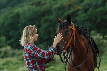 Obraz premium Happy blonde with horse in forest. Woman and a horse walking through the field during the day. Dressed in a plaid shirt and black leggings.