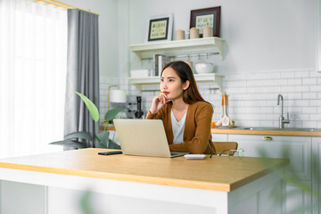 Beautiful young Asian woman working with laptop computer in home kitchen. Work at home. Remote studying, Virtual training, E-learning, Watching online education, Webinar at house