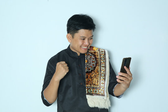 Moslem Asian man excited expression with fist clenched while looking at mobile phone. Wearing a black shirt