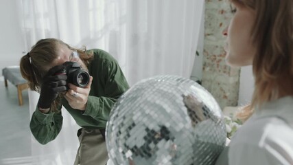 Fashion photographer using digital camera and taking pictures of female model holding disco ball and posing during studio photoshoot. Rack focus