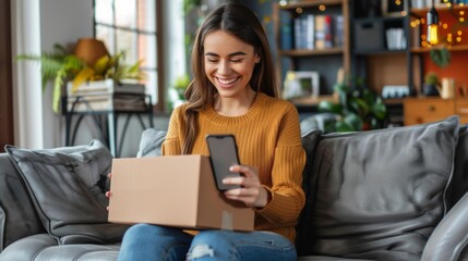 A smiling young woman is sitting on the sofa at home, holding a mobile phone and an open cardboard box in her hand, she is happy about the received package and the purchase.