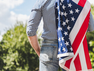 Attractive man in work clothes holding American Flag against background of trees and blue sky. Rear...