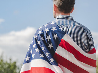 Handsome man holding American Flag against trees and blue sky. View from the back. Close-up,...