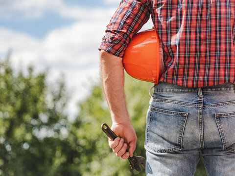 Attractive man in work clothes with a protective helmet against a background of trees and blue sky. Rear view, close-up, outdoors. Labor and employment concept