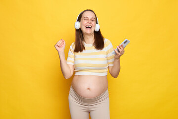 Overjoyed cheerful positive pregnant woman wearing striped T-shirt standing with smartphone being in wireless headphones enjoying favorite soundtracks or radio station