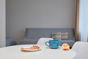 Table in kitchen with cup, apple and slice of pizza. Dinner or snack with hot beverage coffee or tea in mug, copy space for advertisement or promotional concept