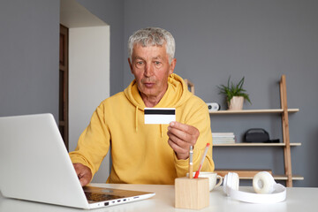 Shocked senior man in yellow hoodie sitting at home workplace with laptop and making inline paying...