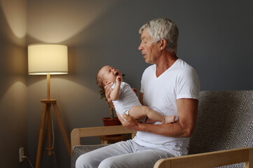 Elderly man sitting indoors with his newborn grandson in his arms enjoying their happy family time...