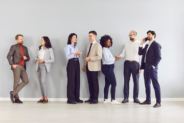 Diverse team of cheerful happy business employees meeting in office, standing at wall, smiling, talking about work, professionals celebrating common success, group portrait, workspace discussion 