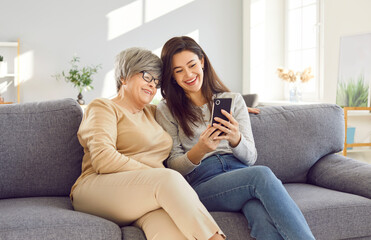 Smiling happy adult woman using mobile phone with cheerful mature senior mother parent sitting on sofa at home browsing apps or having online video call. Family relationship, leisure concept.