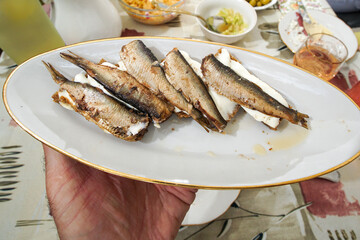 A man's hand holds a saucer containing sandwiches with sprats over a table with treats.
