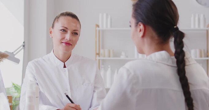 Dermatologist or cosmetologist giving professional skin care consultation. Happy young woman in white uniform holding pen and clipboard and talking to young girl patient in her office