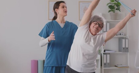 Physical therapist helps elderly woman perform exercises with rubber band to improve flexibility. Smiling retired woman performs side bends under supervision of young female therapist. 4k footage.