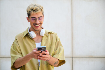 Fototapeta premium A man with a moustache and glasses is smiling while looking at his cell phone. He is wearing headphones and a yellow shirt