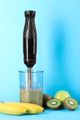 Hand blender with mixture of ingredients and fresh fruits on light blue background