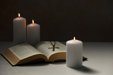 Cross with chain, burning candles and Bible on light table against black background. Religion of...