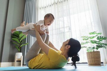 Asian Mother lifting smiling baby during exercise at home. Indoor lifestyle photography with plants in background. Family and fitness concept