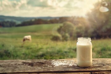 A glass of milk on a wooden table in a field setting with a cow and sunlight in the background