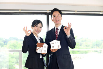 住宅模型を持ちOKポーズをするスーツ姿の男女のビジネスパーソン