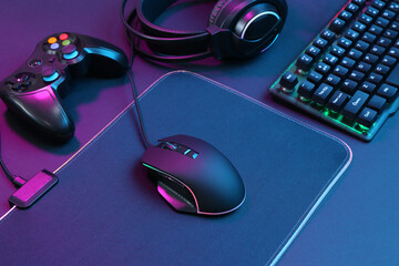 Computer mouse, RGB keyboard, game controller and headset in neon lights on dark background