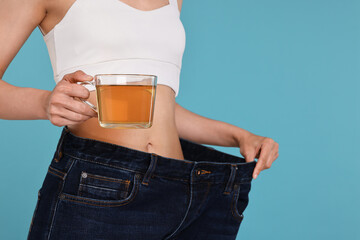 Weight loss concept. Woman with cup of diet tea and big pants on light blue background, closeup