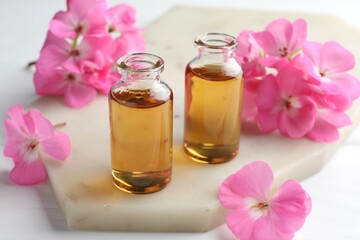 Bottles of geranium essential oil and beautiful flowers on white table, closeup