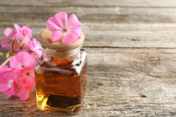 Bottle of geranium essential oil and beautiful flowers on wooden table, closeup. Space for text