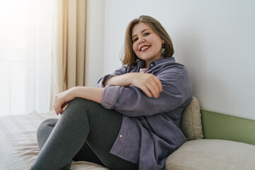 Smiling plus-size woman sitting on bed, looking at camera with relaxed and confident expression. Cozy home environment, self-acceptance and positivity. Casual and comfortable lifestyle.