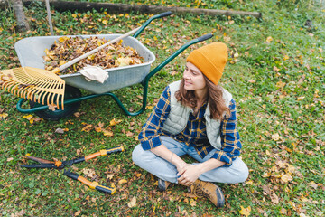 A young woman removes fallen autumn leaves with a rake in the garden of a country house.