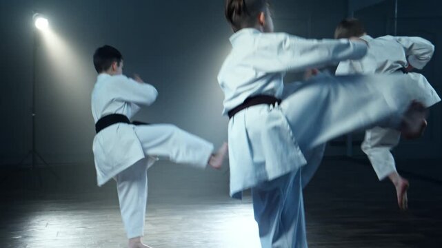 Children in white kimono training karate at smoky background. Three boys in karate class standing side by side with serious expressions. Group perform karate kata. Children synchronously practice