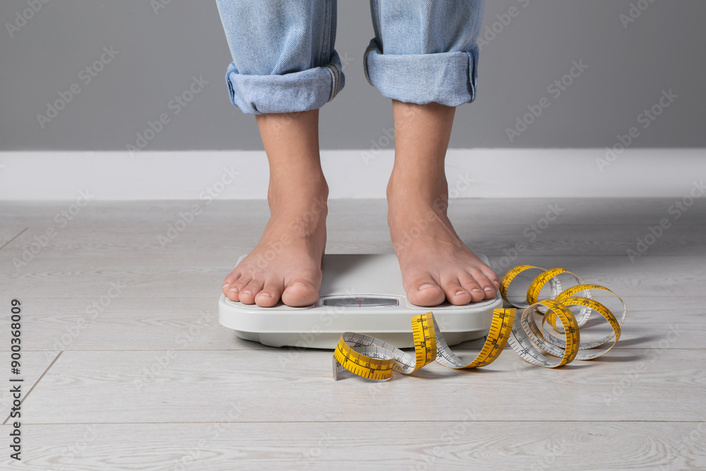 Poster Eating disorder. Woman standing on floor scale and measuring tape indoors, closeup