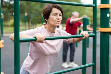 Obraz premium Mature woman doing push-ups from the crossbar on outdoor sports ground