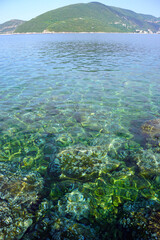 Summer seascape from Luštica peninsula: transparent sea water through which you can see stones with algae on the bottom, and a hill on the Košare peninsula. Beach vacation in the Bay of Kotor