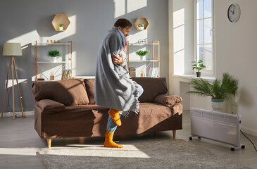 Man freezes inside house or apartment in very cold winter. Man wearing blanket and socks shivering...