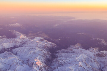 Fototapeta premium View out of an airplane window of a snow-covered mountain range at sunset