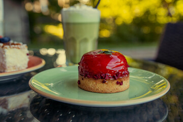 A single raspberry tartlet sits on a green plate, glistening with a red glaze. The background is blurred, suggesting a warm, sunny afternoon with a cool drink waiting to be enjoyed.