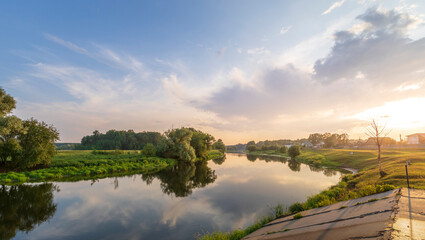 A serene rural river scene with lush trees by its banks. The sky is tinted orange and pink by the setting sun, casting a warm glow on the water