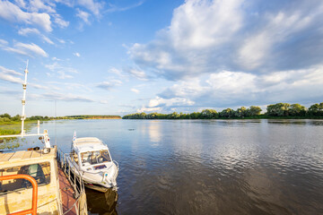 A calm river flows under a sunny sky with fluffy clouds. Two boats are docked on the riverbank, one...