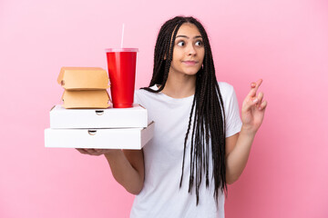 Teenager girl with braids holding pizzas and burgers over isolated pink background with fingers crossing and wishing the best
