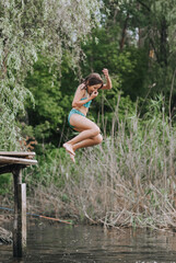 Fototapeta premium Athletic beautiful teenage girl, child jumping from a wooden bridge, pier into the water on a lake outdoors in nature in the summer on vacation. Photography, portrait, childhood concept, sport.