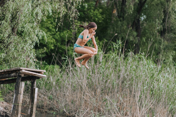Athletic beautiful teenage girl, child jumping from a wooden bridge, pier into the water outdoors...