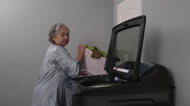Handheld Shot Footage, An elderly woman loads clothes into a top-loading washing machine. She smiles as she closes the machine's lid and adjusts the settings, demonstrating household chores.