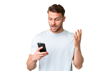 Young handsome caucasian man over isolated chroma key background looking at the camera while using the mobile with surprised expression