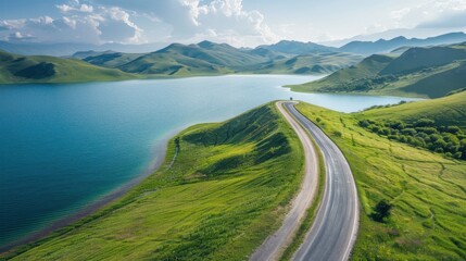 Serpentine Road to Serenity: An aerial vista of a winding road tracing a vibrant green mountain range, bordering a tranquil turquoise lake under a majestic sky. 