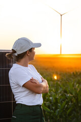 Fototapeta premium Woman wearing white cap stands next to solar panel at sunset. Wind turbines in the background