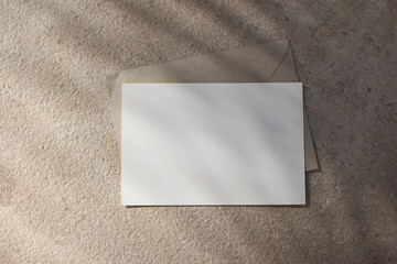 Empty horizontal greeting card invitation mockup, craft envelope. Beige limestone backgound in sunlight. Palm leaf shadow overlay. Modern branding template. Tropical summer, vacation. Flat lay, top